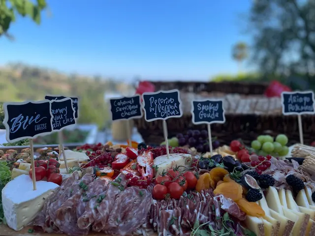 An outdoor charcuterie board with rolled salami, prosciutto, grapes and labelled cheeses against a hillside view