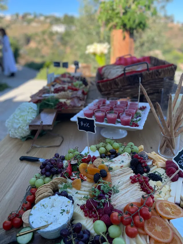 An outdoor buffet: soup shooters on a cake stand, a cheese and fruit board, breadsticks and white hydrangeas