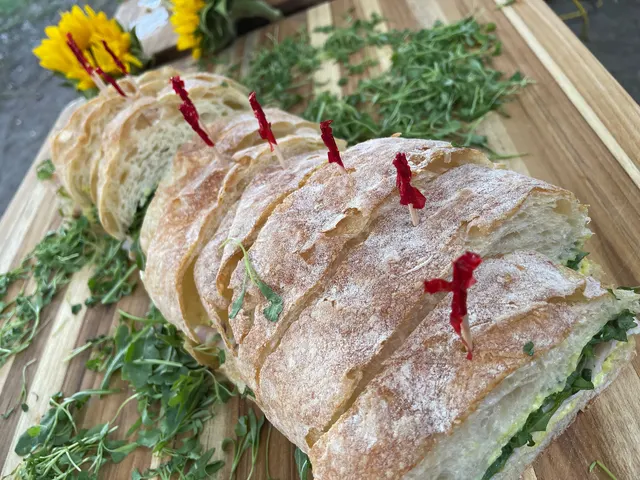 A rustic sandwich loaf sliced into portions with red-tipped picks, on a board scattered with herbs and sunflowers