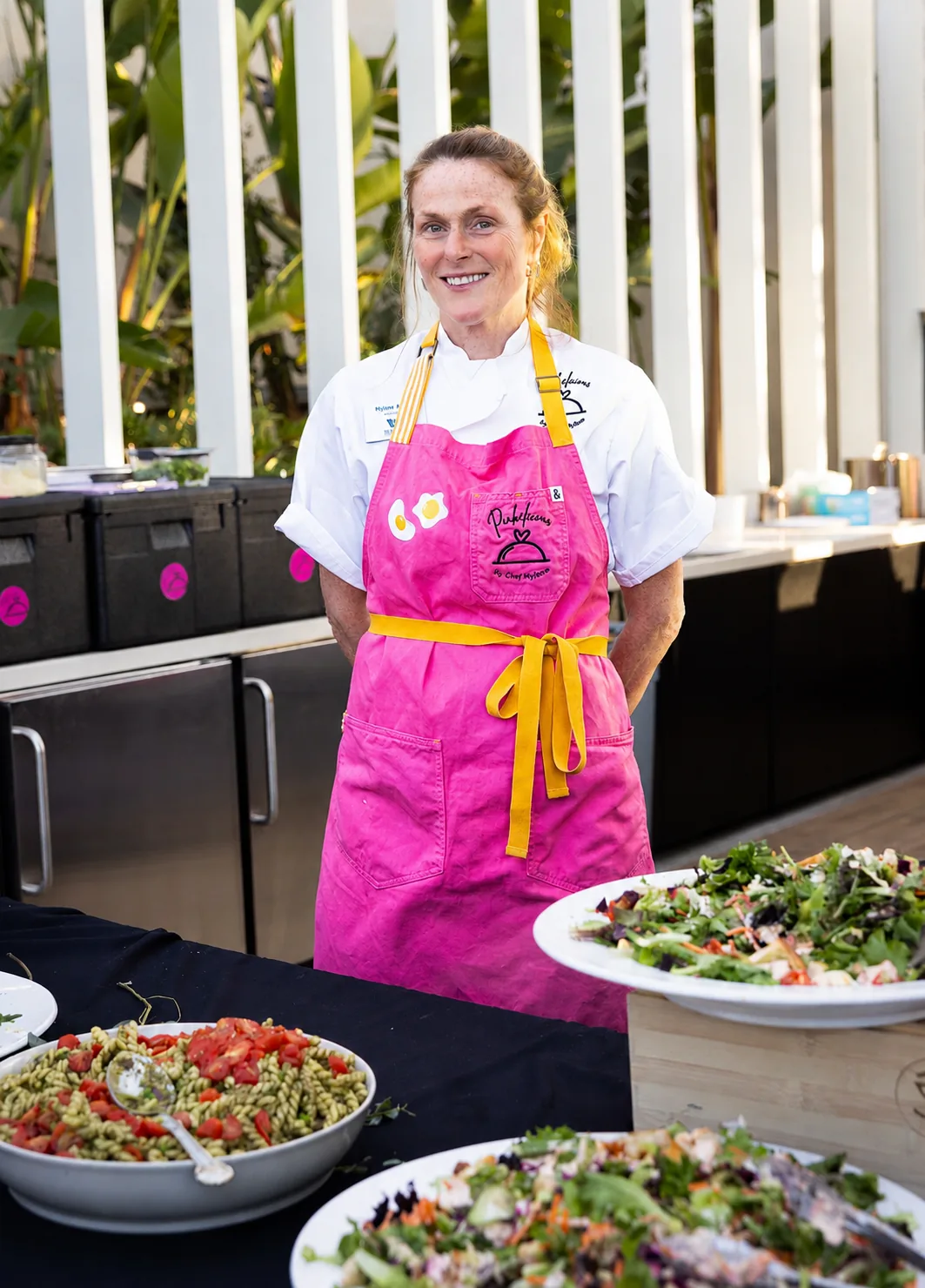 Chef Mylène smiling in her pink Pinkelicious apron beside a large bowl of mixed green salad at an outdoor event
