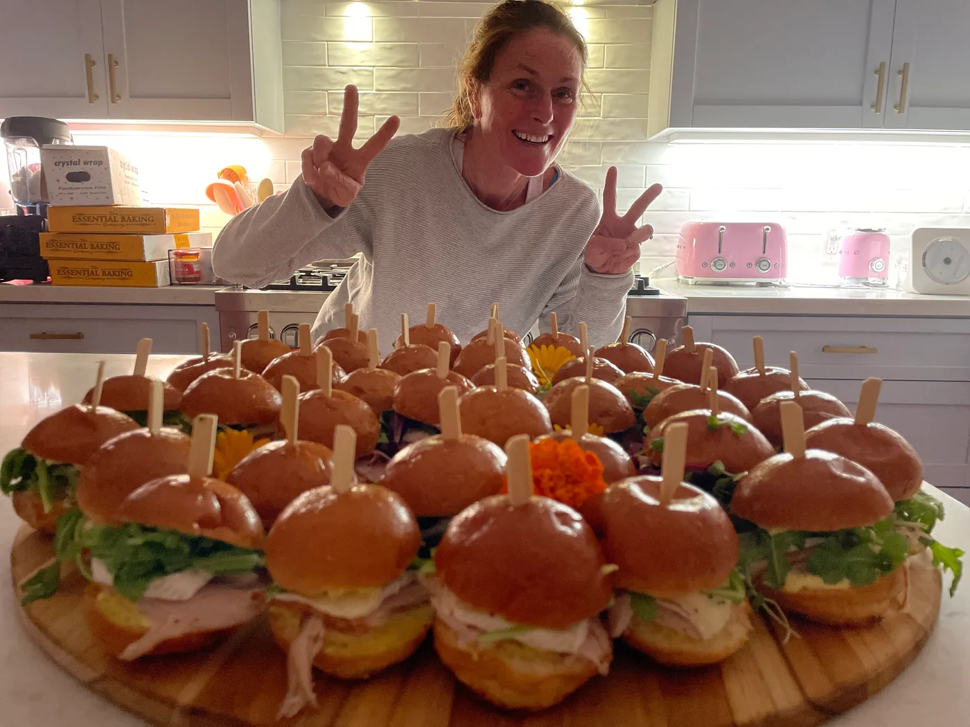 Chef Mylène flashing peace signs in her kitchen behind a wooden board piled with brioche sliders